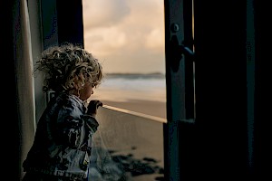 This image shows a small child at a window, family by James Bowden, UK  photographer, represented by Horton-Stephens photographer’s agents specialises in natural and authentic  lifestyle outdoors nature photographic images.