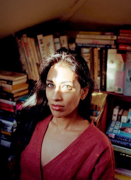 A woman in red top looks up at camera with light on her green eyes from a corner of books portrait by Meghan Eagles Top London Lifestyle Photographer represented by Horton-Stephens Agency